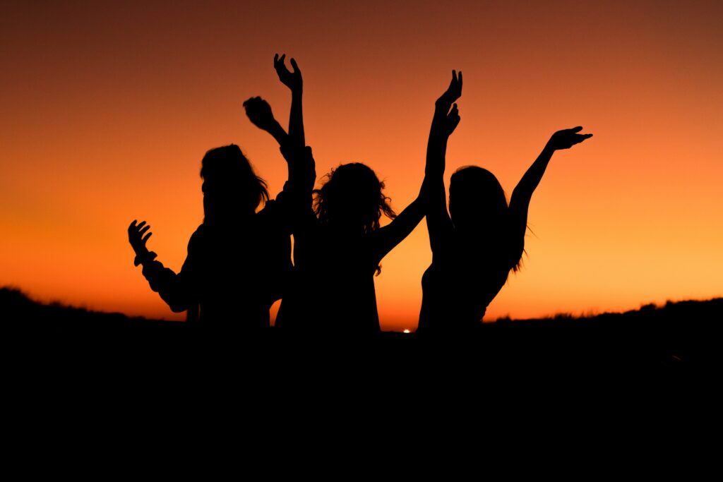 Three dancing women silhouetted against a red and orange sunset, it is unclear if they are celebrating midlife.