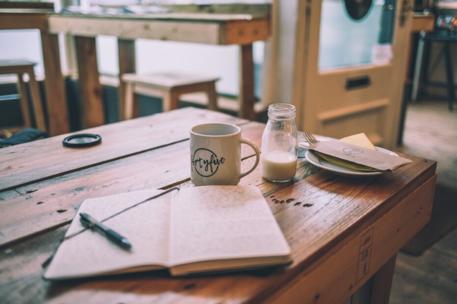 A journal, a pen, a coffee and a pot of milk in a cafe.