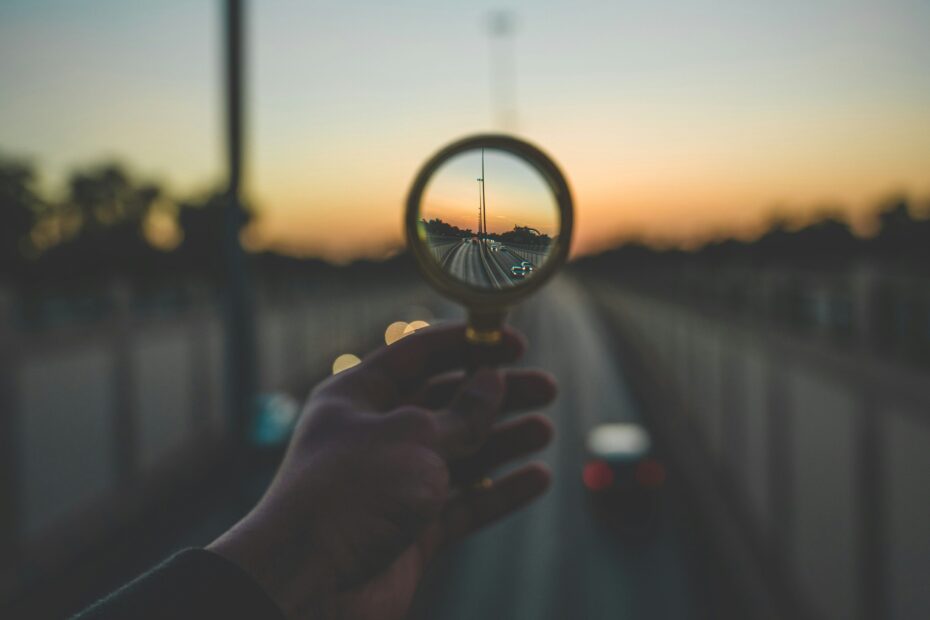 A hand holding a magnifying glass in front of a bridge at sunset. The centre of the magnifying glass is in focus, the rest of the image is blurry.