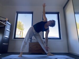 Em on her yoga mat in triangle pose. Feet double-shoulder-width apart, arms open, upper body tilted to her left hand side. She is looking up towards her right hand which is vertical.