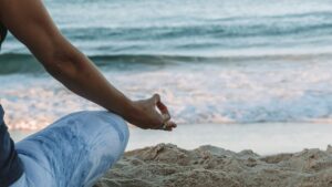 A woman sitting in the lotus position is mostly out of frame. We can see her right leg, arm and hand from behind. She is sitting on the beach with the ocean in the background.