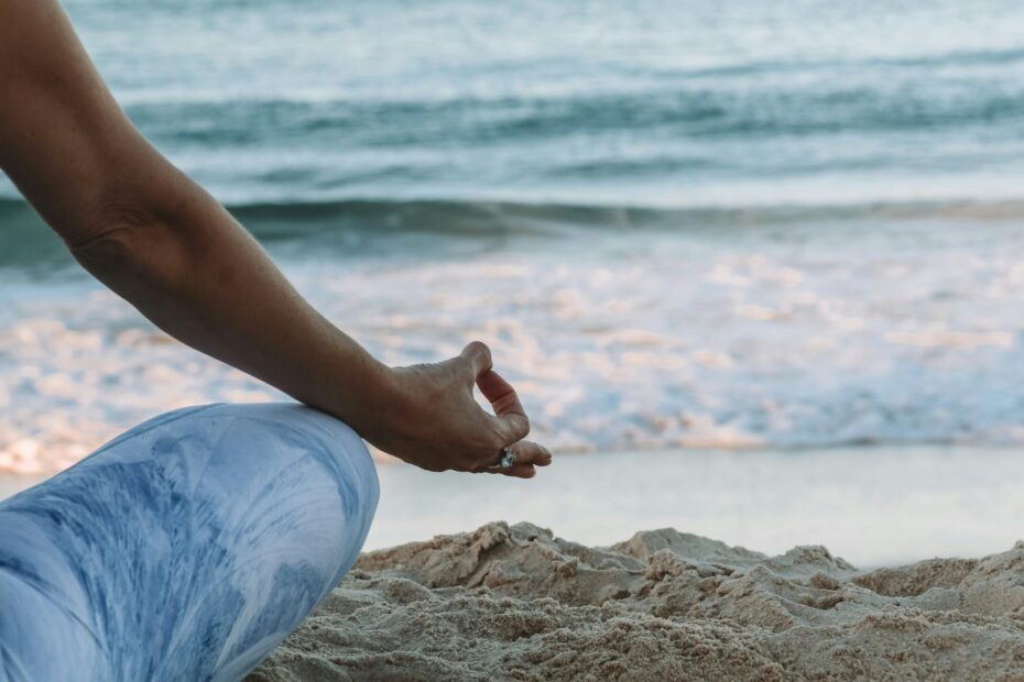 A woman sitting in the lotus position is mostly out of frame. We can see her right leg, arm and hand from behind. She is sitting on the beach with the ocean in the background.