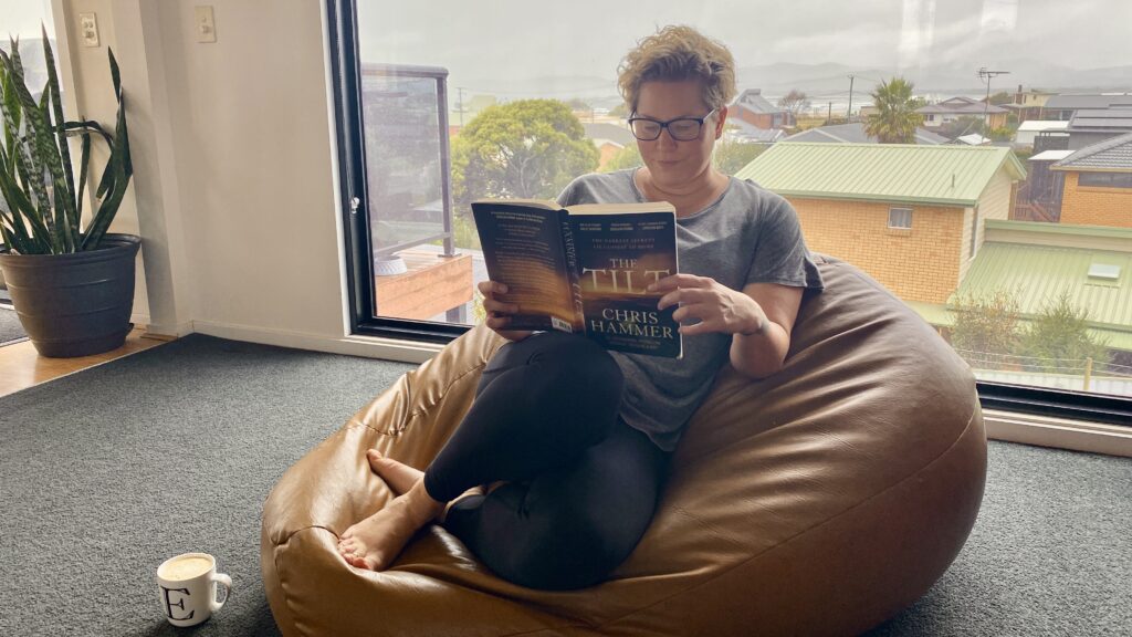 Em sits on a brown leather beanbag (made by her uncle) in her lounge room reading The Tilt by Chris Hammer. She has a white coffee mug with an E on it on the floor beside her. She is dressed for comfort, wearing tights, a grey top and bare feet. She also wears glasses. In the background is a large picture window looking out towards the water. It is a grey, overcast day.