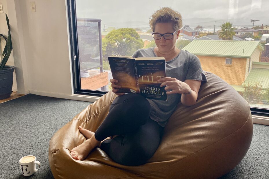 Em sits on a brown leather beanbag (made by her uncle) in her lounge room reading The Tilt by Chris Hammer. She has a white coffee mug with an E on it on the floor beside her. She is dressed for comfort, wearing tights, a grey top and bare feet. She also wears glasses. In the background is a large picture window looking out towards the water. It is a grey, overcast day.