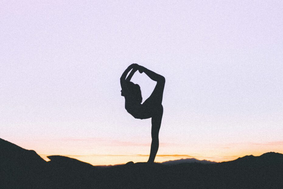 woman silhouetted against a purple and orange sunset stands on one leg with her other leg bent up behind her and over her head. This woman is clearly not Em.